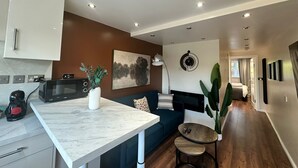 Una cocina moderna con microondas, cafetera y un florero con plantas. Hay una zona de comedor con mesa y sillas, un sofá y una planta grande. El ambiente tiene piso de madera e iluminación empotrada en el techo.