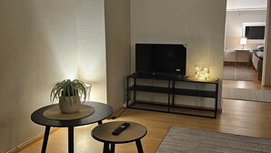 Ein Wohnzimmer mit einer Couch, einem kleinen Tisch mit Pflanze, einem Fernseher auf einem Schrank und einer Lampe.