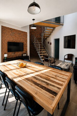 Ein modernes Wohnzimmer mit einem großen Esstisch aus Holz, schwarzen Ledersesseln, einem an einer Ziegelwand montierten Fernseher und einer Treppe, die in ein Obergeschoss führt.