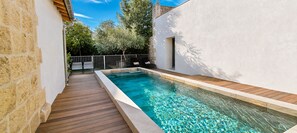Ein Schwimmbecken mit einer Holzterrasse, umgeben von einer weißen Mauer und einer Steinmauer, mit einem Metallzaun und Bäumen im Hintergrund.