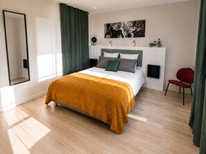 A modern bedroom with a large bed, a headboard, a mounted artwork, a side table, a red chair, and a mirror.
