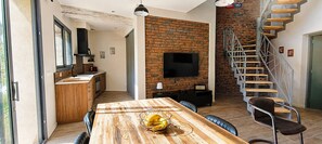 Una cocina moderna con pared de ladrillo, una zona de comedor con mesa de madera y una escalera con barandilla de metal.