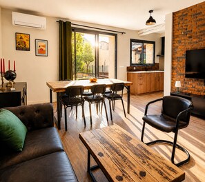 Una sala moderna con un sofá, una mesa de centro de madera y un comedor con sillas. Se observa una cocina con un televisor y una pared de ladrillo.