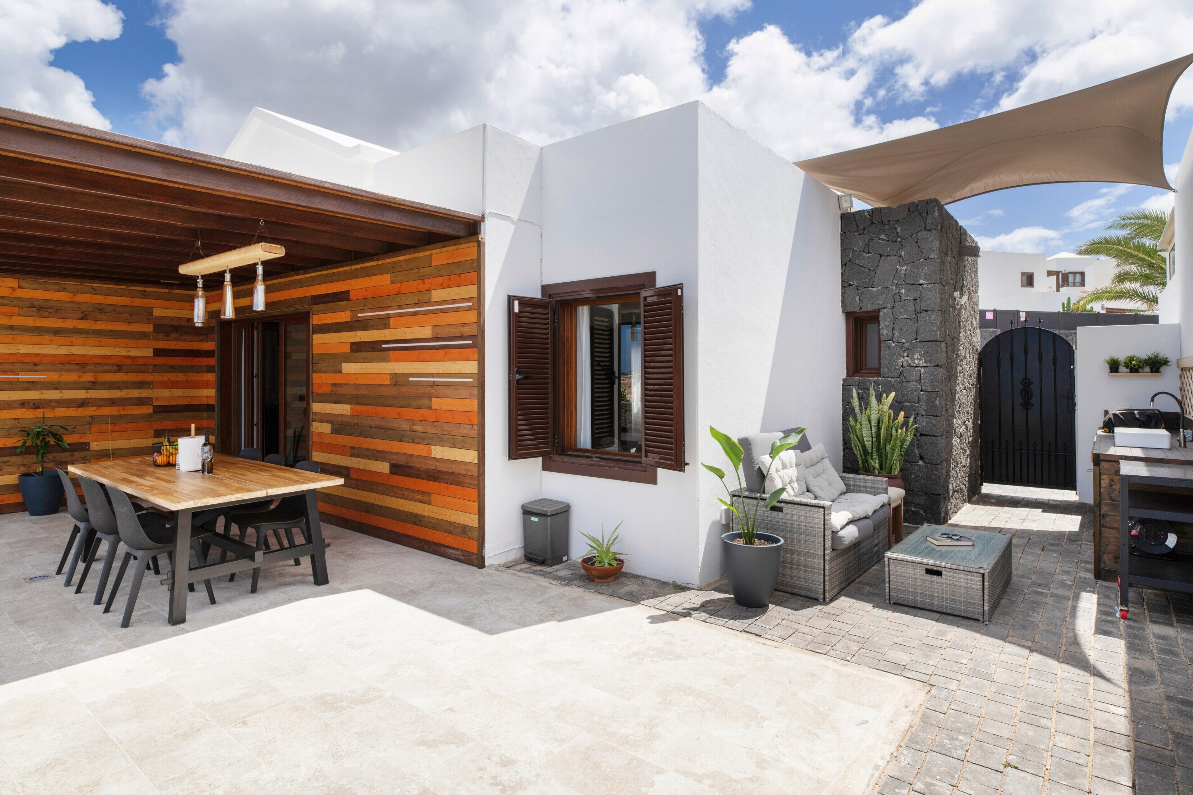 A modern outdoor patio with a wooden table and chairs, a stone wall, and a black gate.