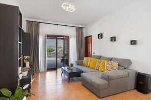 Ein Wohnzimmer mit grauem Sofa, Couchtisch und TV-Schrank. Eine Glastür öffnet sich zu einem Außenbereich.