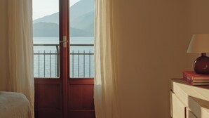 A room with a view of a lake and mountains, a wooden door, a bedside table with a lamp and books, and a bed. - Authentic house with beautiful view of lake como (Musso)