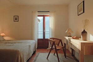 Una habitación con cama, una silla, un escritorio, una lámpara, un florero y una puerta que da a un balcón.