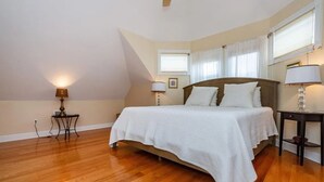 Een slaapkamer met een houten vloer, een bed met wit beddengoed, twee bedlampjes, een raam met gordijnen en een klein tafeltje met een lamp.