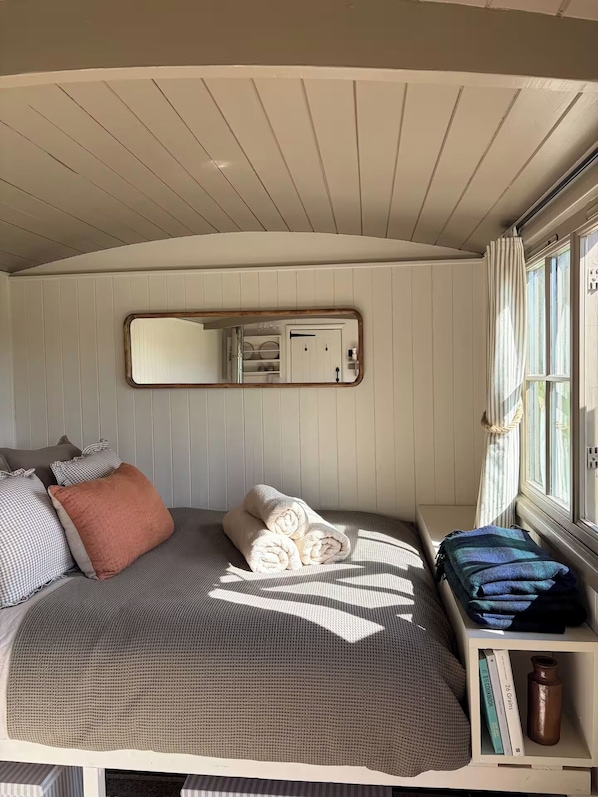 A cozy bedroom with a bed, pillows, a rolled-up towel, a shelf with books and a water bottle, a window with curtains, and a mirror on the wall.