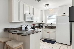 Eine Küche mit weißen Schränken, einer Mikrowelle, einem Herd und einem Kühlschrank. Zwei Hocker und ein Fenster mit Jalousien sind ebenfalls vorhanden.