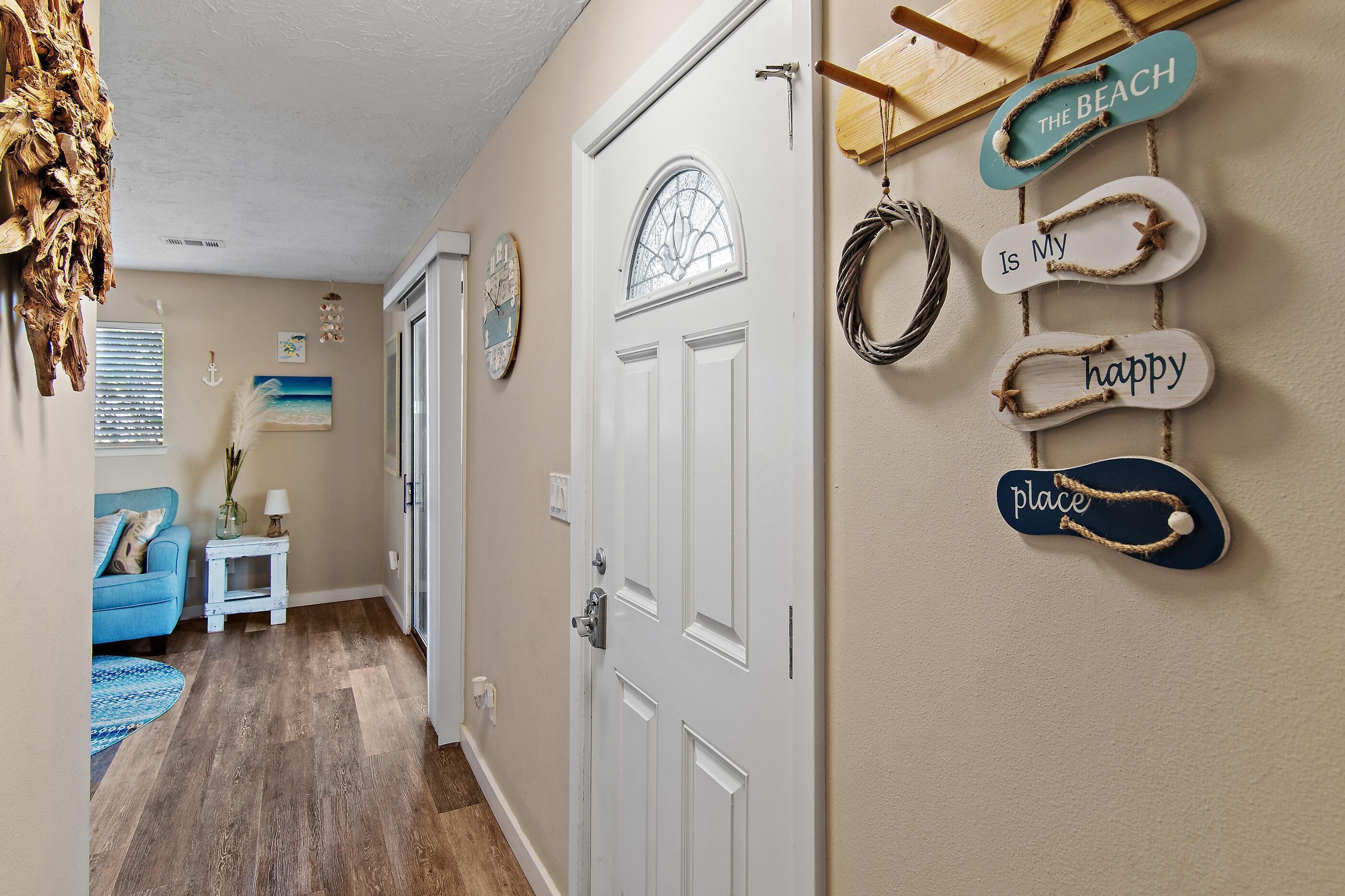 A hallway with a wooden floor, a white door, and wall decorations including a sign that says 'The Beach is My happy place.'
