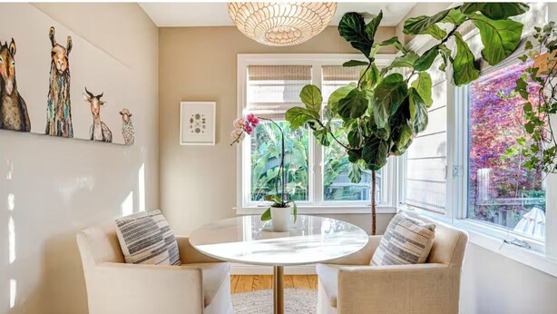 A room with a round table, two chairs, a large potted plant, wall art, and a window with a view of greenery. - Downtown Mill Valley Vintage Spanish Villa with Beautiful Garden (Mill Valley)