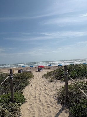 Ein Sandweg am Strand, der zum Meer führt, mit Liegestühlen und Sonnenschirmen.