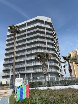 Ein mehrstöckiges Hotel mit Balkonen, Palmen und einem Schild, das auf einen Strandbereich hinweist.