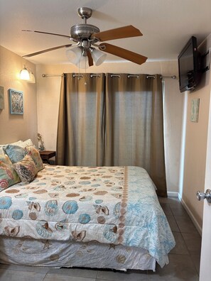Ein Schlafzimmer mit Deckenventilator, Vorhängen, einem Bett mit gemustertem Überwurf, einem Nachttisch und einem an der Wand befestigten Fernseher.