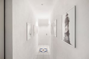 Un couloir aux murs ornés de tableaux encadrés, doté d’un sol carrelé et terminé par une porte blanche.