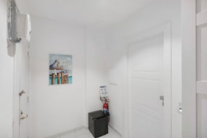 Un couloir aux murs blancs, une porte, un petit réfrigérateur noir, un extincteur et un tableau représentant des chevaux.