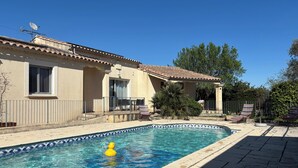 Una piscina con borde de azulejos, un patito de goma amarillo y una casa de techo con tejas y paredes color crema.