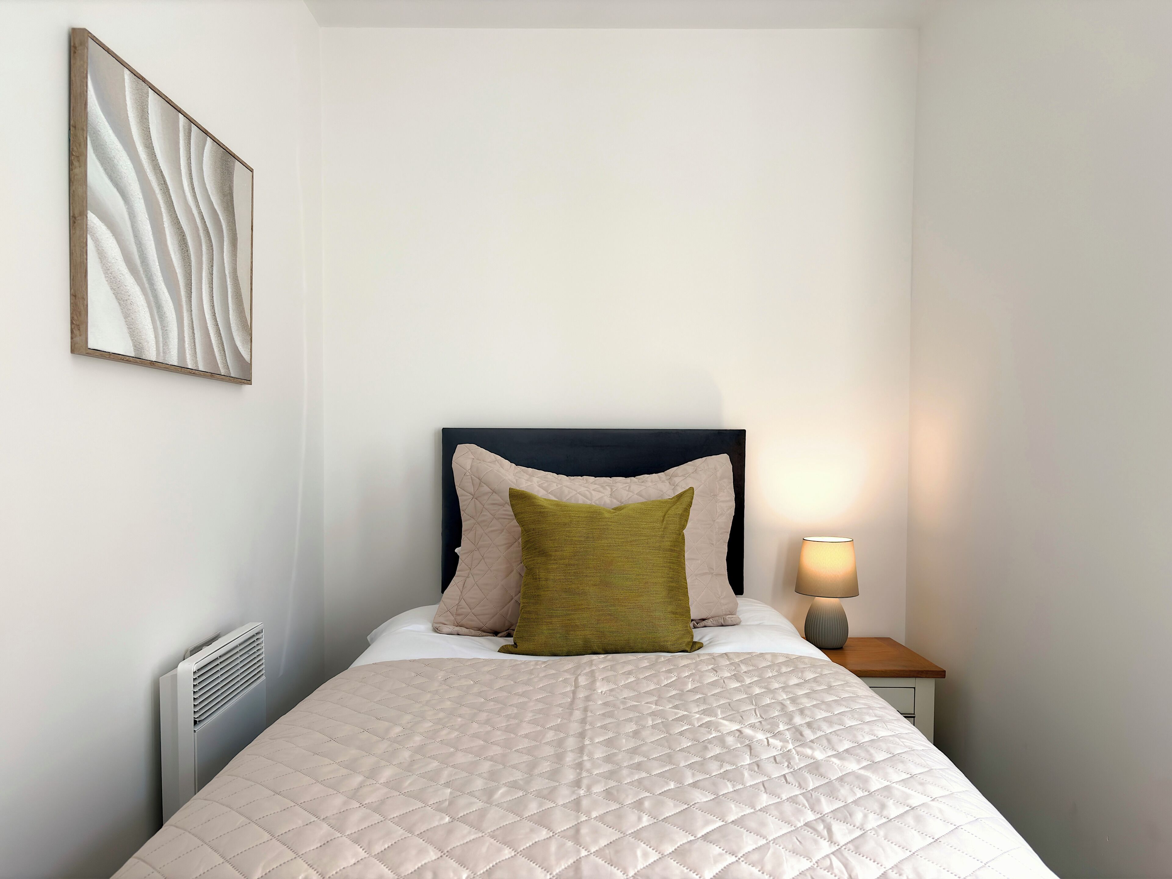 A neatly made bed with a quilted cover, a bedside lamp, and a radiator.