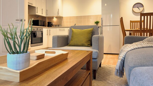 A compact kitchen with white cabinets, a microwave, and a refrigerator. There is a grey sofa with a green pillow and a wooden table with a plant. - St Johns Courtyard Suite, Bridgnorth with free private parking (Bridgnorth)