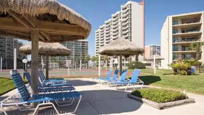 A resort area with lounge chairs, thatched umbrellas, and modern buildings in the background. - Beachfront Saida condo with Gulf views, resort pools, room for 6 and a $75 beach gear rental credit. (South Padre Island)