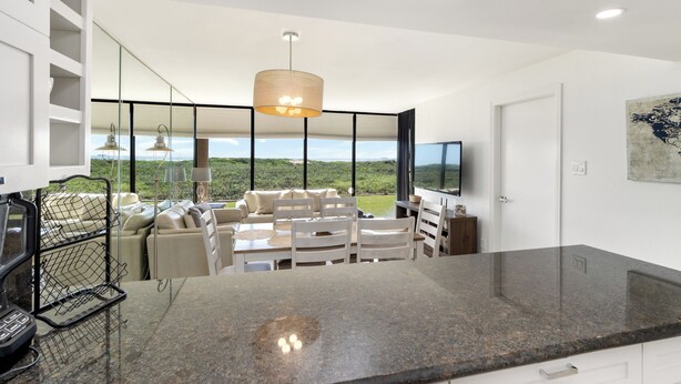 Eine moderne Küche mit Granit Arbeitsplatte, ein Essbereich mit Tisch und Stühlen, ein großes Fenster mit Blick auf die Landschaft und ein an der Wand befestigter Fernseher.