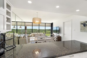 Cozinha moderna com bancada de granito, área de jantar com mesa e cadeiras, janela grande com vista para a paisagem e televisão na parede.