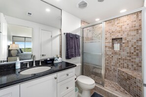 A bathroom with a glass-enclosed shower, a white toilet, a sink with a black countertop, and a mirror. - 3BR Beachfront Condo at Sea Vista with Ocean Views (South Padre Island)