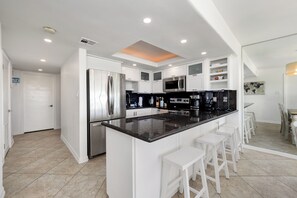 A modern kitchen with white cabinetry, a central island with bar stools, stainless steel appliances, and recessed ceiling lighting. - First-floor beachfront Sea Vista condo with Gulf views, pools + $75 beach gear rental credit (South Padre Island)