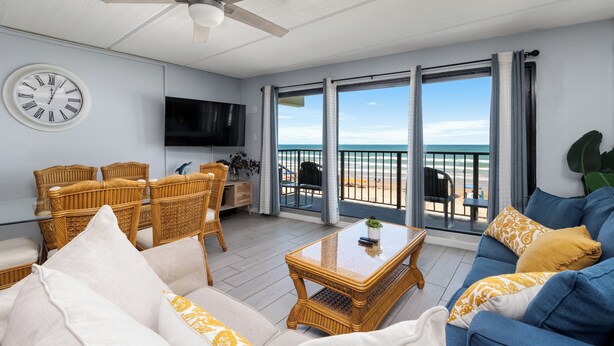 Ein Wohnzimmer mit Deckenventilator, einer runden Wanduhr, einem Flachbildfernseher, Korbmöbeln, einem blauen Sofa und einem Balkon mit Blick auf den Strand.