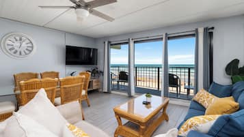 A living room with a ceiling fan, a round wall clock, a flat-screen TV, wicker furniture, a blue sofa, and a balcony with a view of the beach.