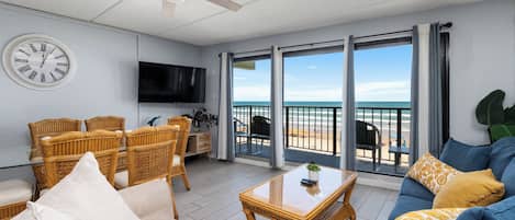 Ein Wohnzimmer mit Deckenventilator, einer runden Wanduhr, einem Flachbildfernseher, Korbmöbeln, einem blauen Sofa und einem Balkon mit Blick auf den Strand.