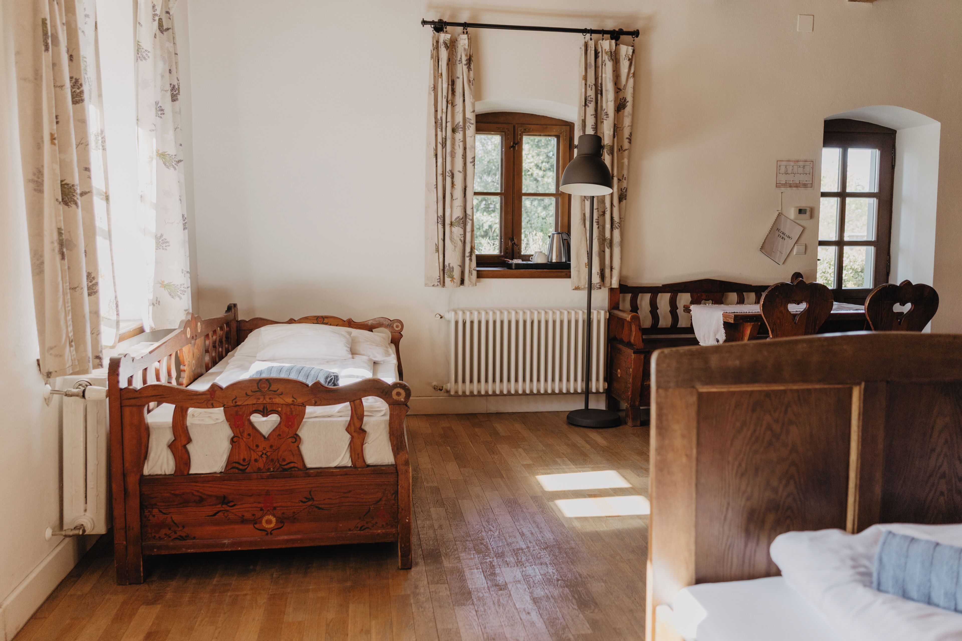 A room with a wooden bed, a desk with a chair, a radiator, and a window with curtains.