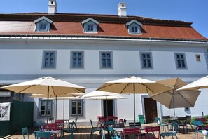 Un bâtiment avec un toit recouvert de tuiles rouges, des murs blancs et de nombreuses fenêtres. On y trouve des tables et des chaises à l’extérieur, ainsi que des parasols.
