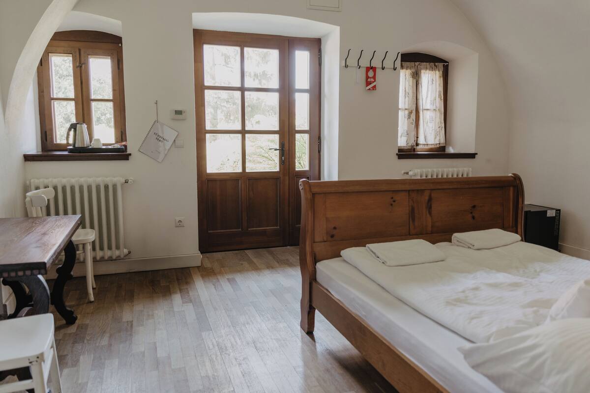 A bedroom with a wooden bed, a desk, a chair, a radiator, and two windows.