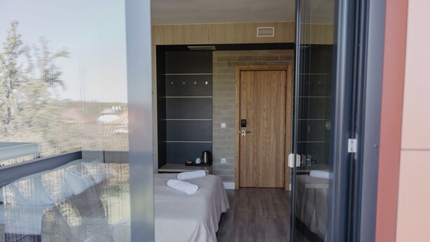 A modern hotel room with a balcony, a wooden door, and a bed with a white bedspread.