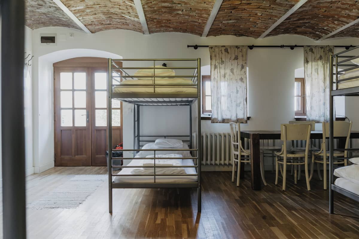 A dormitory room with bunk beds, a wooden floor, a radiator, and a window with curtains.