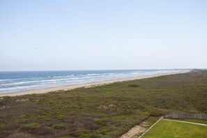 A beach with a grassy area, a wooden fence, and a wooden structure in the distance. - Oceanfront Sea Vista condo with wraparound balcony, pools + $75 beach gear rental credit (South Padre Island)