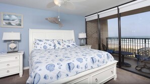 Quarto com cama, ventilador de teto, mesa de cabeceira, luminária, quadro e vista para a praia através de uma varanda.