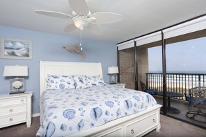 A bedroom with a bed, a ceiling fan, a nightstand, a lamp, a framed picture, and a view of the beach through a balcony. - Oceanfront Sea Vista condo with wraparound balcony, pools + $75 beach gear rental credit (South Padre Island)