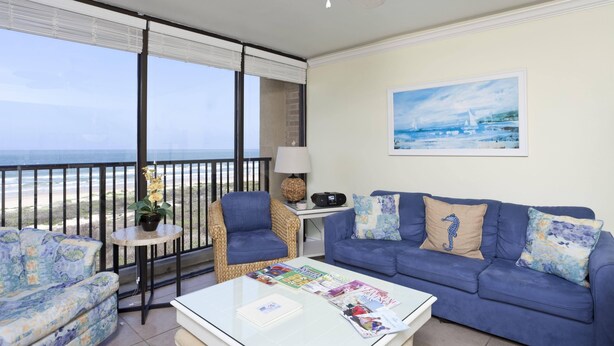 Uma varanda com vista para o mar, um sofá azul, uma cadeira de vime e uma pequena mesa com abajur e rádio.