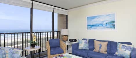 Uma varanda com vista para o mar, um sofá azul, uma cadeira de vime e uma pequena mesa com abajur e rádio.