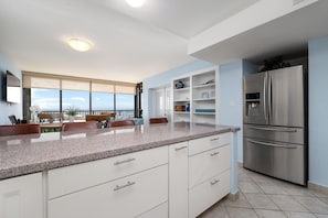 A modern kitchen with a large island, white cabinetry, a stainless steel refrigerator, and a dining area with a view. - Beachfront condo with Gulf views, 2 pools, hot tub & private beach access! (South Padre Island)