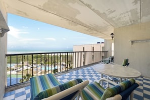 A balcony with a checkered floor, a table, and chairs, overlooking a pool and beach area. - Spacious beachfront corner condo with Gulf views, 3 pools & hot tubs! (South Padre Island)