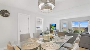 A modern living room with a glass dining table, grey sofas, a TV, and a view of the outdoors. - First-floor Edgewater condo with direct beach access, Gulf views + $75 beach gear rental credit (South Padre Island)