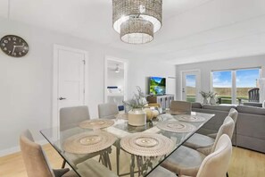 A modern living room with a glass dining table, grey sofas, a TV, and a view of the outdoors. - First-floor Edgewater condo with direct beach access, Gulf views + $75 beach gear rental credit (South Padre Island)