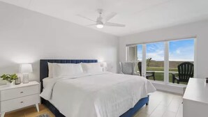 A bedroom with a bed, nightstands, a desk, a chair, a ceiling fan, and a large window with a view. - First-floor Edgewater condo with direct beach access, Gulf views + $75 beach gear rental credit (South Padre Island)