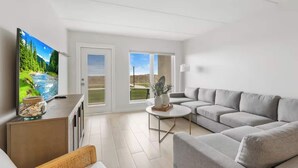 A modern living room with a grey sofa, a TV on a wooden stand, a small table with decorative items, and a view of a green landscape through the sliding doors. - First-floor Edgewater condo with direct beach access, Gulf views + $75 beach gear rental credit (South Padre Island)