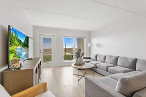 A modern living room with a grey sofa, a TV on a wooden stand, a small table with decorative items, and a view of a green landscape through the sliding doors. - First-floor Edgewater condo with direct beach access, Gulf views + $75 beach gear rental credit (South Padre Island)