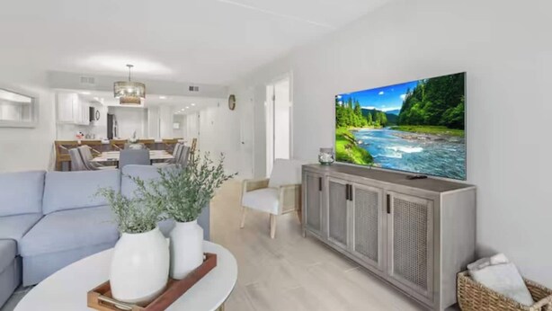 A modern living room with a flat-screen TV, a sofa, a dining area, and a kitchen in the background. - First-floor Edgewater condo with direct beach access, Gulf views + $75 beach gear rental credit (South Padre Island)